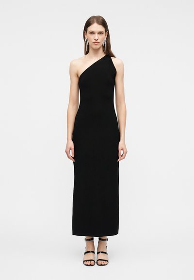 Mulher em pé, vestindo um vestido preto longo de um ombro só, saltos pretos com tiras e brincos longos pendurados, contra um fundo claro e simples.