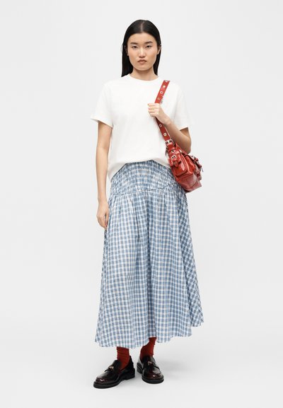 Femme portant une chemise blanche à manches courtes, une longue jupe à carreaux bleue et blanche, des chaussettes rouges, des mocassins noirs, et portant un sac à bandoulière en cuir rouge.