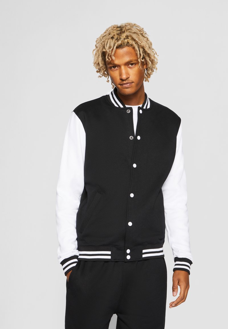 Chaqueta varsity en blanco y negro con cierre de botones, puños acanalados y cuello a rayas, que presenta una textura suave y dos bolsillos frontales.