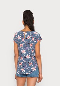 Blusa corta de manga corta azul con flores rosas y blancas y hojas verdes, escote redondeado y detalle de ojal en la parte posterior.