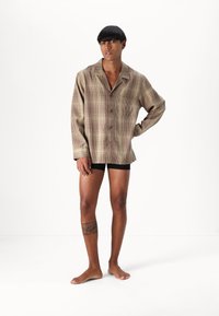 Randig skjorta i beige och brunt, lätt tyg, avslappnad passform, långa ärmar, en ficka. Bärs över svarta shorts.
