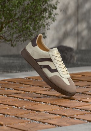 Zapatilla crema y marrón con cordones, punta de ante y suela marrón oscuro flotando sobre suelo de baldosas de madera al aire libre.