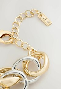 Bracciale in metallo oro e argento con grandi anelli intrecciati e una chiusura, dotato di un piccolo ciondolo rettangolare con una singola gemma.