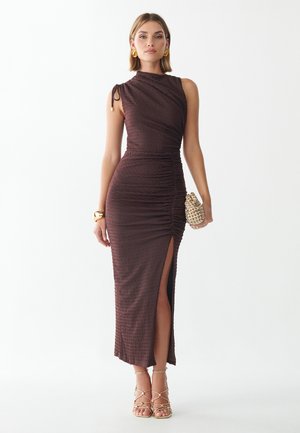 Mujer con un vestido oscuro marrón sin mangas fruncido con abertura alta, pendientes dorados, pulsera, sandalias y sosteniendo un bolso de mano tejido.