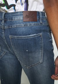 Vue arrière d'une personne portant un jean bleu délavé avec un patch en cuir « Kings will Dream » ainsi qu'une partie d'une chemise multicolore rentrée à l'intérieur.