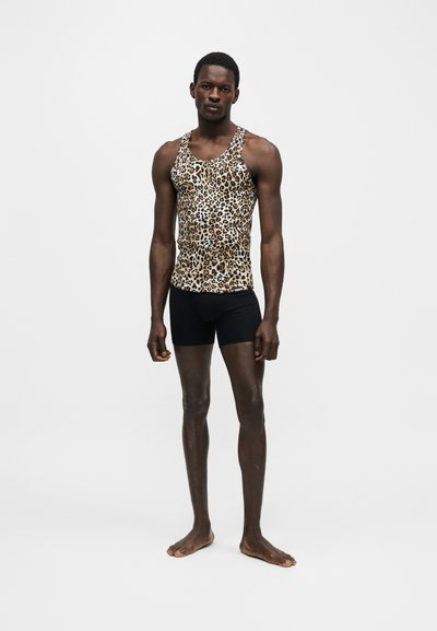 Ψηλός άντρας που στέκεται ξυπόλυτος, φοράει αμάνικο τοπ με σχέδιο λεοπάρδαλης και στενά μαύρα σορτς σε απλό λευκό φόντο.