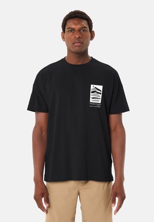 Uomo che indossa una maglietta nera con una grafica astratta bianca e il testo "INSIEME alla scoperta di nuovi sentieri nella natura", abbinata a pantaloni beige.