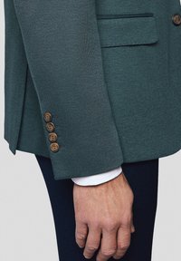 Zielony teksturowany blezer o dopasowanym kroju, cztery brązowe guziki na mankiecie oraz widoczny biały mankiet koszuli pod spodem.