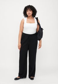 Top sans manches blanc à encolure carrée, pantalon noir à jambes larges et chaussures plates noires. Sac noir avec une sangle froncée. Cheveux bouclés, arrière-plan neutre.