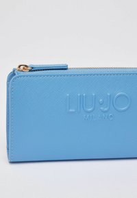 Portefeuille bleu clair texturé avec logo embossé "LIU·JO MILANO", fermeture éclair dorée et languette. Présente une forme rectangulaire avec des bords lisses.