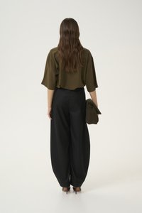 Mujer de espaldas vistiendo un top ancho oliva, pantalones negros de pierna ancha, tacones altos, sosteniendo un bolso de mano a juego en color oliva.