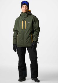 Grön vattentät jacka med orange detaljer, fickor med dragkedja och justerbar huva, kombinerad med svarta byxor och handskar, lämplig för vintersport.