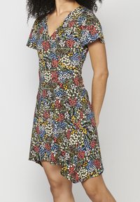 Femme portant une robe à manches courtes et col en V avec un motif floral coloré en rouge, jaune, bleu et blanc sur fond noir.