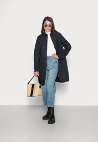 Lungo cappotto navy trapuntato con colletto alto, indossato sopra un dolcevita bianco. Abbinato a jeans chiari a vita alta e stivaletti neri alla caviglia. Tiene una borsa intrecciata.