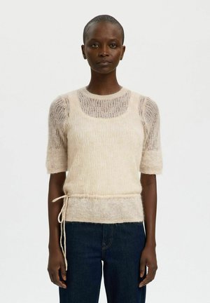 Vrouw met kort haar draagt crèmekleurige, pluizige, gebreide lagen tops en donkere jeans, staand tegen een eenvoudige witte achtergrond.
