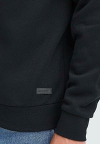 Sort sweatshirt med tekstureret stof, ribbede manchetter og kanten, samt et lille vævet mærke på den nederste venstre side. Båret over blå denim.