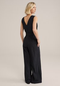 Tuta nera senza maniche con una profonda scollatura a V sul retro, vita evidenziata da una cintura, gambe larghe e tessuto texturizzato. Design semplice con una silhouette pulita.