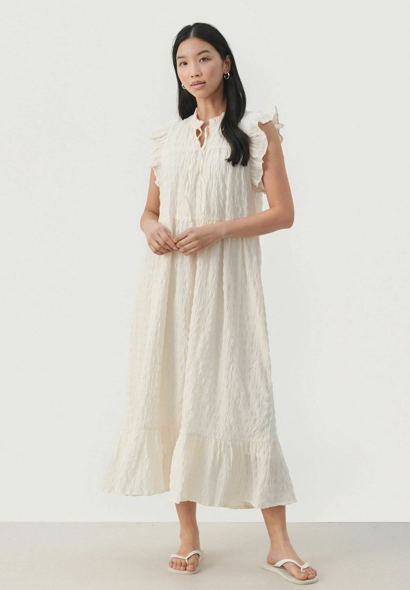 Robe maxi blanche texturée avec des manches capes à volants, détail de boutons et ourlet en couches. Associée à des sandales blanches à retour.