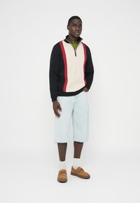 Gebreide trui met rits in zwart, crème en rood met een gestructureerd patroon; gecombineerd met lichte denim shorts en bruine schoenen.