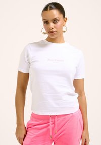 Balta trumpa rankovėmis marškinėliai, pagaminti iš medvilnės, su šviesiai rožiniu "Juicy Couture Sport" logotipu ant krūtinės, derinami su rožinėmis sportinėmis kelnėmis.
