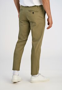 Slanke olijfgroene broek met een gladde textuur, twee achterzakken en zwarte knopen. Gecombineerd met witte sneakers en sokken.