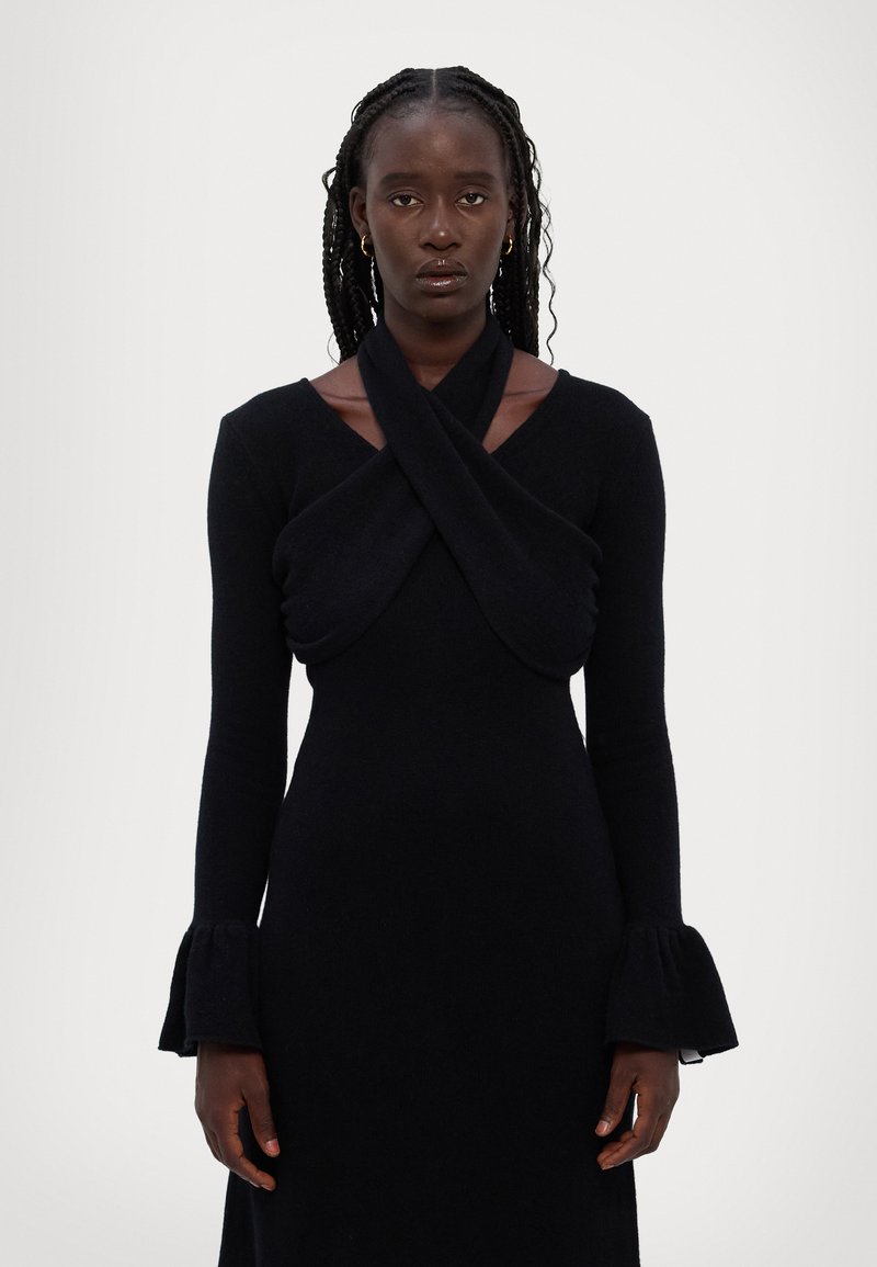 Abito nero a maniche lunghe con scollatura torcigliata e polsini svasati, realizzato in un tessuto morbido e texturizzato. Design minimalista con finitura liscia.