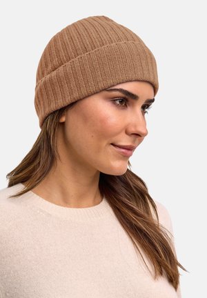 Donna con berretto di maglia beige e maglione color crema, mostrata di profilo su uno sfondo chiaro uniforme.