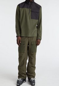 Olijfgroene fleece trui met een zwart bovenpaneel en een borstzak. Gecombineerd met bijpassende cargobroek met grote zakken. Zwarte skischoenen.