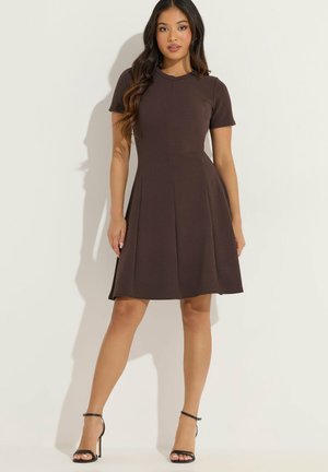 Kvinde står iført en kortærmet, knælængde mørkebrun kjole med sorte ankelrem høje hæle mod en ensfarvet baggrund.