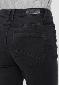 Los jeans de mezclilla negra presentan un acabado texturizado, bolsillos traseros prominentes y una etiqueta de cuero con logo en la cintura.
