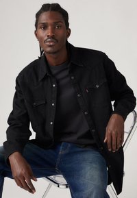 Camisa preta de denim com botões de pressão, dois bolsos no peito e costura contrastante. Usada por cima de uma camisa escura, combinada com jeans azuis.