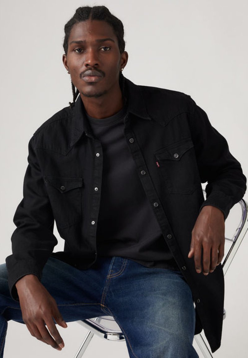 Camisa preta de denim com botões de pressão, dois bolsos no peito e costura contrastante. Usada por cima de uma camisa escura, combinada com jeans azuis.