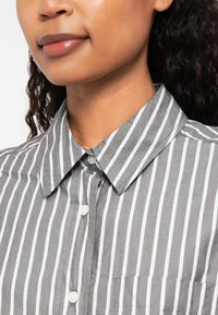 Camicia grigio striata con strisce verticali bianche, caratterizzata da un colletto e un'apertura con bottoni. Include un dettaglio con taschino sul lato sinistro.