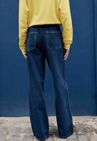 Vaqueros anchos de pierna ancha en azul oscuro con bolsillos traseros y costuras visibles, combinados con una camisa de manga larga de color amarillo brillante.