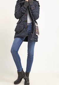 Mörkblå parka med knappstängning och fickor, lagd över en grå topp. Åtsittande blå jeans och svarta ankelstövlar med struktur på sulorna.