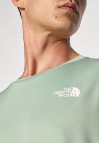 Camiseta de manga corta verde claro, hecha de un tejido texturizado, con un logo blanco de "The North Face" en el lado izquierdo del pecho.