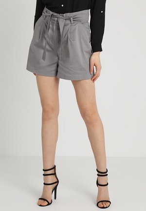 Femme portant un short paperbag gris taille haute avec ceinture nouée, un haut noir à manches longues et des sandales noires à talons hauts et à lanières.