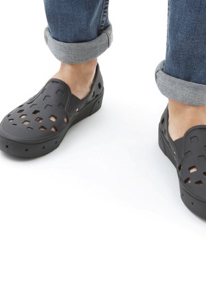 Fødder iført sorte slip-on sko med ventilationshuller, kombineret med opkrogede blå jeans mod en hvid baggrund.