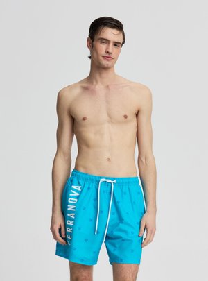 Giovane uomo con capelli scuri che indossa pantaloncini da bagno blu brillante con scritta bianca "TERRANOVA" e un laccio bianco, in piedi davanti a uno sfondo semplice.
