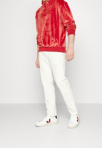 Sudadera de terciopelo rojo con cordón, pantalones blancos y zapatillas blancas con acentos en burdeos y logo. Conjunto casual que resalta la textura y el color.