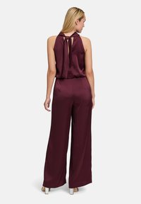 Burgundy jumpsuit in satin fabric, featuring a halter neck, open back with tie detail, and wide legs. Smooth texture and fluid drape.