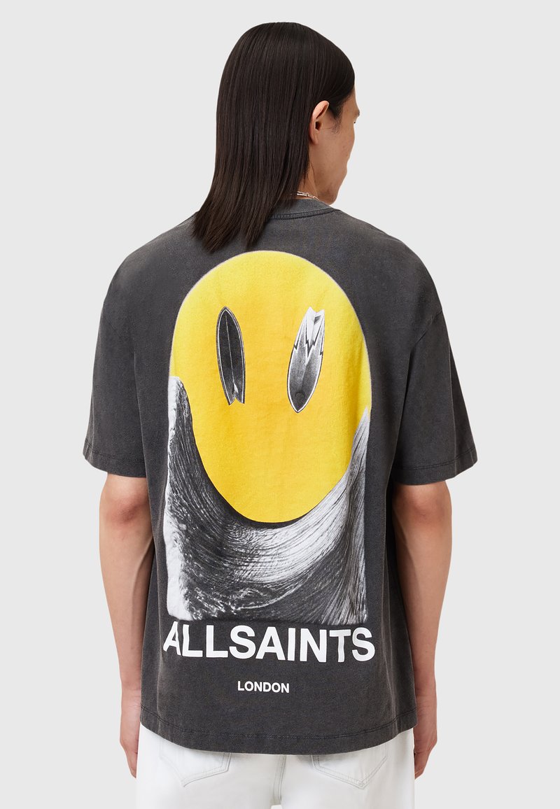 Grå bomulds T-shirt med et stort gult smiley ansigt grafik på ryggen, med sorte detaljer og teksten "ALLSAINTS LONDON" nederst.