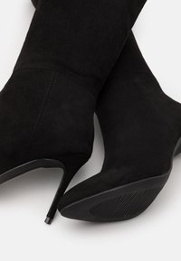 Bottes à talons hauts en suède noir avec un bout pointu, une finition texturée et un talon fin. La semelle présente un motif côtelé pour une meilleure adhérence.