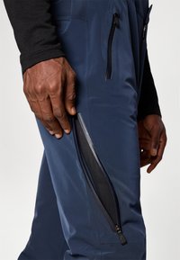 Pantaloni ușori, de culoare bleumarin, prezintă o buzunar cu fermoar pe coapsă și o textură netedă. Manșetele sunt strâmte pentru un aspect aerodinamic.