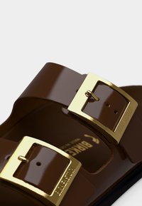 Κοντινό πλάνο στα καφέ λουριά σανδαλιών με αγκράφες σε χρυσαφί απόχρωση και ορατή ανάγλυφη επιγραφή "BIRKENSTOCK" στο μεταλλικό εξάρτημα.