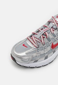 Silver athletic shoe with mesh upper, red accents, and reflective details. Features gray straps, textured toe cap, and black outsole.