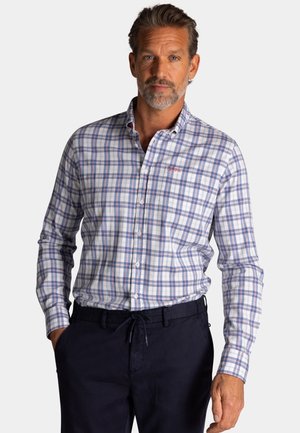 Man met grijs haar en baard, gekleed in een blauw-wit geruit overhemd en donkere broek, staand met handen in de zakken.