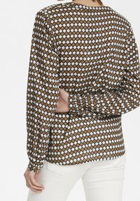 Femme portant un chemisier à manches longues marron et blanc à motifs losanges, avec un pantalon blanc, un bras plié derrière le dos.