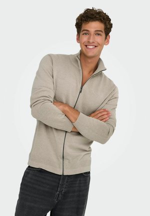 Lichtbeige ritsvest met textuurpatronen, ribgebreide mouwen en zoom, en een hoge kraag, gecombineerd met donkere jeans.