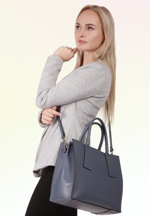 Blond vrouw in een grijze blazer die een grote donkerblauwe leren handtas over haar schouder draagt en opzij kijkt.